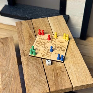 Könnte beinhalten: Ein Miniatur-Brettspiel aus Holz mit roten, gelben, grünen und blauen Spielfiguren. Das Spielbrett hat eingravierten Text und liegt auf einer Holzoberfläche. Ein weißer Würfel ist ebenfalls vorhanden.