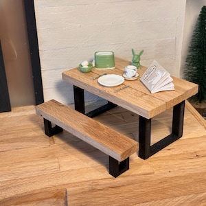 Peut inclure: Une table de pique-nique miniature en bois avec un cadre en m&eacute;tal noir et un banc assorti. La table est dress&eacute;e avec une assiette blanche, une tasse et une soucoupe, un petit r&eacute;cipient vert et un livre intitul&eacute; "The Little Farmhouse".