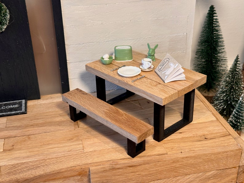 Peut inclure: Une table de pique-nique miniature en bois avec un cadre en m&eacute;tal noir et un banc assorti. La table est dress&eacute;e avec une assiette blanche, une tasse et une soucoupe, un petit r&eacute;cipient vert et un livre intitul&eacute; "The Little Farmhouse".