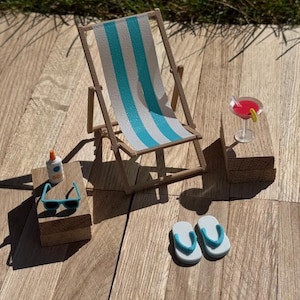 May include: Miniature beach scene with a striped blue and white deck chair, sunglasses, sunscreen, a cocktail, and flip-flops. The scene is set on a wooden surface, evoking a relaxing summer vibe.