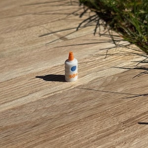 May include: A miniature white bottle with an orange cap and a blue circle with a white dot. The bottle has the number "50+" printed on it. The bottle is sitting on a wooden surface.