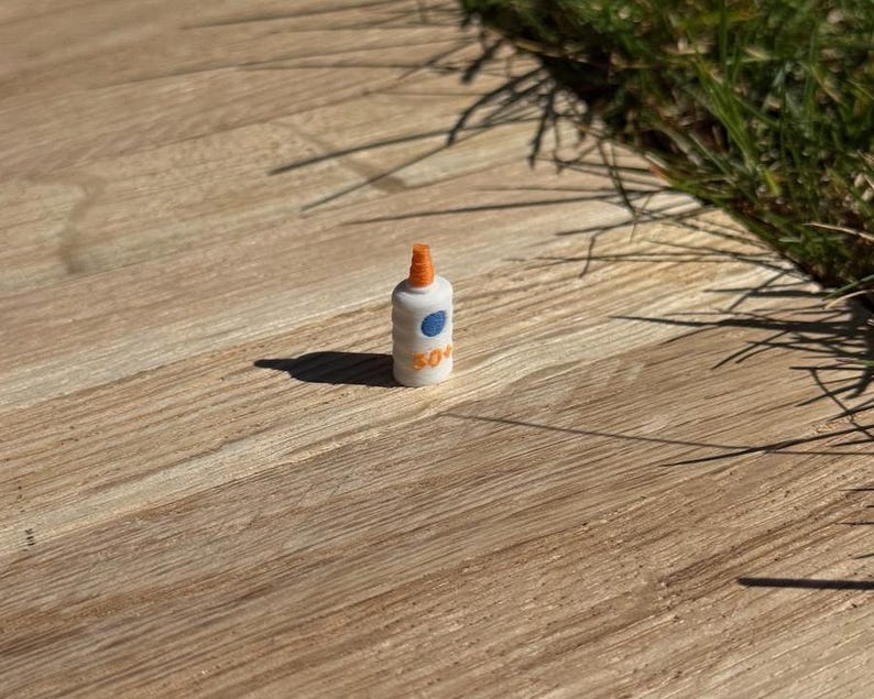 May include: A miniature white bottle with an orange cap and a blue circle with a white dot. The bottle has the number "50+" printed on it. The bottle is sitting on a wooden surface.