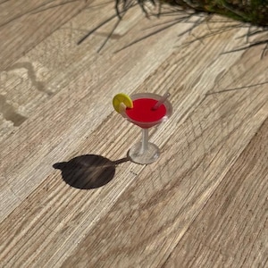 May include: A miniature cocktail in a clear glass with a red liquid, a lemon slice, and a small straw. The drink sits on a wooden surface, casting a shadow.