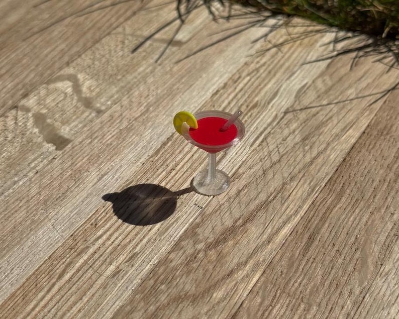 May include: A miniature cocktail in a clear glass with a red liquid, a lemon slice, and a small straw. The drink sits on a wooden surface, casting a shadow.