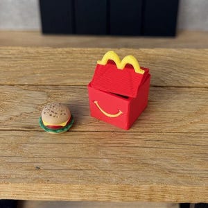 Könnte beinhalten: Eine rote Miniatur-Kunststoffbox mit gelbem Griff und gelbem lächelnden Mund-Design, begleitet von einer kleinen, detaillierten Hamburger-Figur mit beigem Brötchen, roter Tomate, gelbem Käse und grünen Salatblättern.