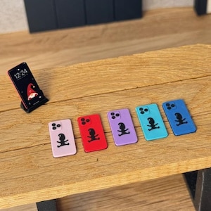 Könnte beinhalten: Eine Sammlung von sechs Miniatur-Spielzeug-Smartphones in verschiedenen Farben wie Hellrosa, Rot, Lila, Hellblau und Dunkelblau. Jedes Gerät zeigt auf der Rückseite die schwarze Silhouette eines Gnoms und eine Dreifachkamera.