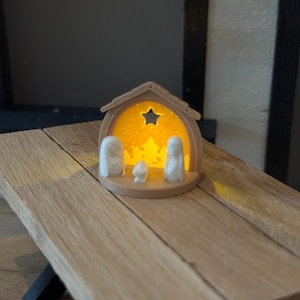 May include: A miniature nativity scene with a wooden frame and a warm, glowing interior. The scene features three white figures and a star-shaped cutout. The wooden structure sits on a wooden surface.