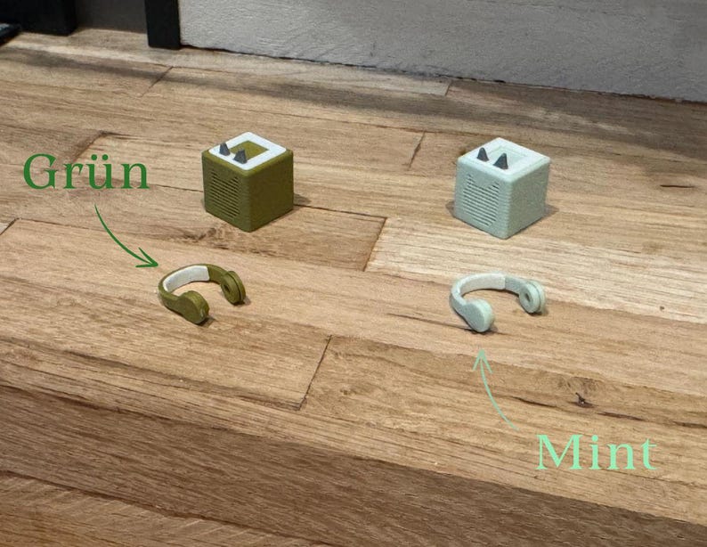 May include: Two small, cube-shaped speakers, one green and one mint green, with headphones next to each. The green speaker has the word "Gr&uuml;n" written above it, and the mint green speaker has the word "Mint" written below it.