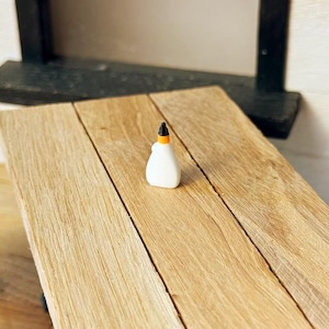 May include: A miniature white glue bottle with an orange and black cap sits on a light brown wooden table. The table is made of planks and is part of a dollhouse or miniature scene. The background includes a black shelf and white wall.