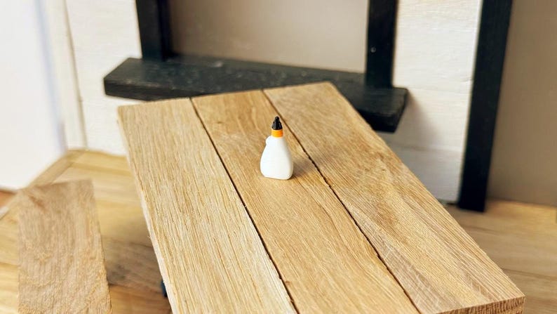 May include: A miniature white glue bottle with an orange and black cap sits on a light brown wooden table. The table is made of planks and is part of a dollhouse or miniature scene. The background includes a black shelf and white wall.
