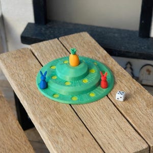 Op de afbeelding: Een miniatuur bordspel met een groene cirkelvormige basis, een wortel en twee konijnenbeeldjes. Een witte dobbelsteen met zwarte stippen ligt op een houten tafel. Het spel is ontworpen voor spelen op kleine schaal.