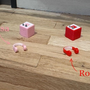 May include: Two small speakers, one pink and one red, with matching headphones. The pink speaker has the word "Rosa" written above it, and the red speaker has the word "Rot" written below it.