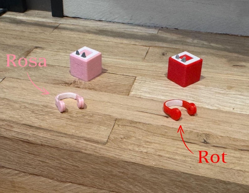 May include: Two small speakers, one pink and one red, with matching headphones. The pink speaker has the word "Rosa" written above it, and the red speaker has the word "Rot" written below it.