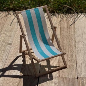 May include: A miniature wooden deck chair with a blue and white striped fabric seat. The chair is set on a wooden surface and casts a shadow. The chair is a decorative item.