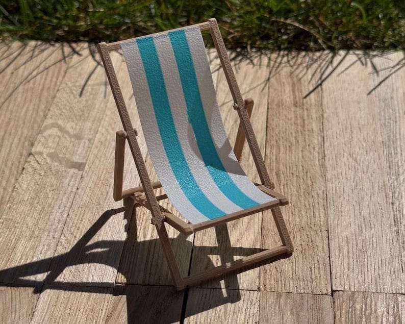 May include: A miniature wooden deck chair with a blue and white striped fabric seat. The chair is set on a wooden surface and casts a shadow. The chair is a decorative item.
