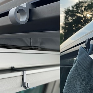 Könnte beinhalten: Zwei schwarze Plastikhaken zum Aufhängen von Gegenständen in einem Auto. Die Haken sind am Dach des Autos befestigt und haben ein schlichtes Design. Ein Haken hängt ein graues Handtuch.