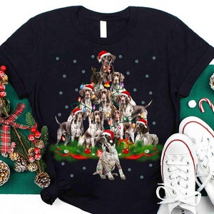 May include: A black t-shirt with a Christmas tree design featuring a group of German Shorthaired Pointer dogs wearing Santa hats. The tree is decorated with red and green ornaments and lights.