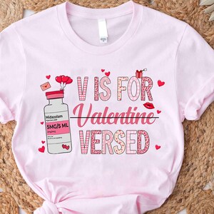 May include: A light pink t-shirt with the text "V IS FOR VALENTINE VERSED" in pink and red lettering. The design includes a cartoon vial of Midazolam and Valentine's Day themed graphics such as hearts and a lipstick kiss.