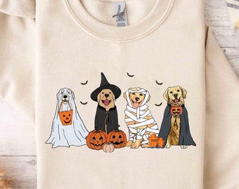 May include: A beige sweatshirt featuring a design of four dogs dressed in Halloween costumes. The dogs are wearing a ghost costume, a witch costume, a mummy costume, and a vampire costume. The dogs are holding pumpkins and bats are flying around them.