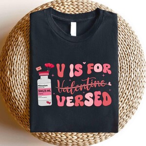 May include: Black t-shirt with the phrase "U is for Valentine VerSed" in pink and red lettering. A cartoon image of a medicine bottle with the label "Midazolam" is on the left side of the shirt. The shirt is on a woven surface.