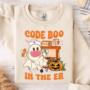 May include: Cream-colored sweatshirt with a Halloween-themed graphic. The design features a ghost wearing a nurse's hat and face mask, holding medical supplies, and a jack-o'-lantern. Text reads "CODE BOO IN THE ER" in orange.