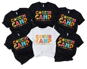 May include: Five short-sleeved t-shirts, four black and one white, each featuring a colorful graphic print that reads COUSIN CAMP 2026. The design includes icons of a sun, guitar, campfire, and tent integrated into the bold, multi-colored lettering.