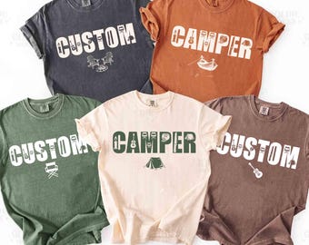May include: Five short-sleeved crew neck t-shirts in charcoal, rust, olive green, cream, and brown. Each shirt features the word CUSTOM or CAMPER in large block letters, with small camping-themed icons integrated into the text and below it.