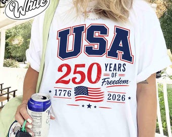 May include: A white t-shirt featuring a graphic with the text USA, 250 YEARS OF FREEDOM, 1776, and 2026 in navy blue and red. The design includes a small American flag illustration, stars, and horizontal lines. A Comfort Color logo is in the corner.