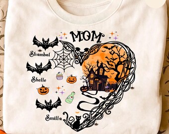 May include: A cream-colored crewneck t-shirt featuring a graphic of a heart-shaped design. The design includes the text MOM, three bats with the names Shambol, Shelle, and Smilla, a spiderweb, pumpkins, a potion bottle, and a haunted house scene.