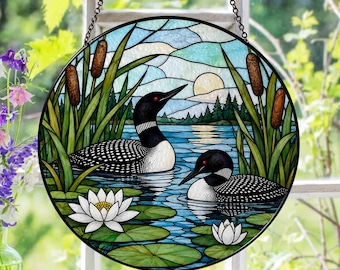 May include: A circular stained glass suncatcher featuring two black and white loons swimming on a blue lake. The scene includes green cattails, lily pads, white water lilies, a distant forest, and a pale sun in the sky, all framed by a black border.