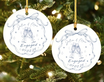 May include: Two white ceramic ornaments with gold hanging strings. Each ornament features a blue ribbon design with the names "Julia & Max", two champagne flutes, and the word "Engaged" with the date "07.02.25".