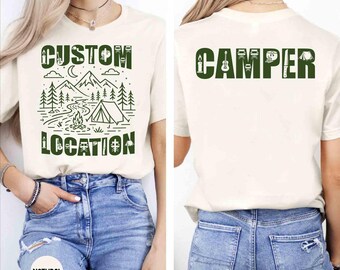 May include: A cream-colored t-shirt featuring a dark green graphic. The front displays a mountain landscape with a tent, campfire, and the text CUSTOM LOCATION. The back features the word CAMPER in large green letters with small icons.