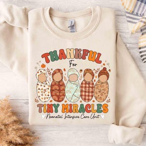 May include: Cream-colored sweatshirt with the words "THANKFUL For TINY MIRACLES Neonatal Intensive Care Unit" in a colorful, retro font. The design features five swaddled babies in various fall-themed patterns.