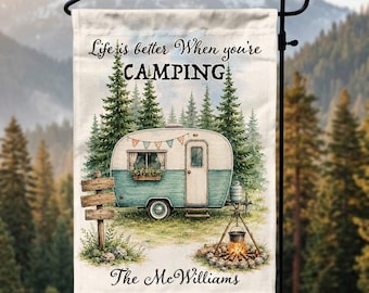 May include: A rectangular garden flag featuring a teal and white vintage camper, a campfire with a hanging pot, and tall pine trees. The text reads Life is better When you're CAMPING and The McWilliams. The flag hangs on a black metal stand.