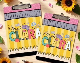 May include: Two rectangular clipboards featuring a yellow pencil-themed design with a pink eraser top, a grey metal ferrule, and a black tip. The text Mrs. CLARA is printed in large, colorful letters, decorated with small icons of books, apples, and pencils.