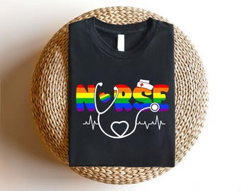 Può includere: Maglietta nera con il testo "NURSE" arcobaleno, uno stetoscopio e una grafica a forma di cuore. Il testo è in uno schema di colori arcobaleno.