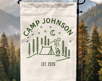 May include: A cream-colored garden flag featuring a dark green line art design of a tent, a campfire, pine trees, stars, and a crescent moon. The text reads CAMP JOHNSON and EST. 2026 in a simple sans-serif font, displayed on a black metal stand.