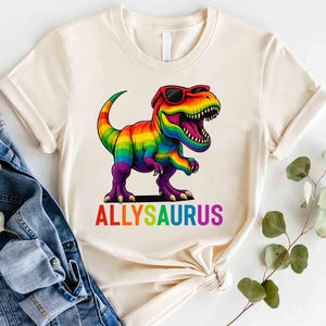 May include: A white t-shirt with a rainbow dinosaur wearing sunglasses and the text "ALLYSAURUS" printed on the front.