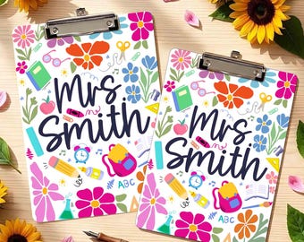 May include: Two white clipboards featuring the text Mrs. Smith in dark blue script, surrounded by colorful illustrations of flowers, a backpack, a pencil, a book, scissors, a clock, and school supplies, with a wooden pen placed between them.