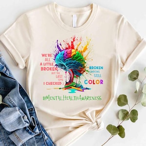 May include: A beige t-shirt with a colorful brain graphic and the text "We're all a little broken but the last time I checked: Broken crayons still color #MentalHealthAwareness".