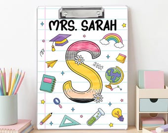 May include: A white clipboard featuring a lined paper design with the text MRS. SARAH at the top. The center displays a large letter S designed like a pencil, surrounded by colorful school-themed illustrations including a globe, rainbow, and books.