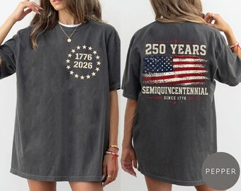 May include: Dark gray t-shirt with a distressed design. The front features the text "1776 2026" surrounded by stars. The back displays an American flag graphic with "250 YEARS SEMIQUINCENTENNIAL SINCE 1776" in red and white.