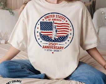 May include: Ivory-colored t-shirt featuring a distressed circular graphic commemorating the 250th anniversary of the United States. The design includes the American flag, the Statue of Liberty, and the text "UNITED STATES OF AMERICA" and "250th ANNIVERSARY 1776-2026".