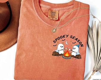 May include: A terracotta-colored t-shirt featuring a graphic of two ghosts roasting marshmallows over a campfire on a log. The text "SPOOKY SEASON" is printed above the ghosts, accompanied by small stars, a crescent moon, and bat silhouettes.