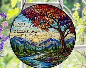 May include: A circular stained glass suncatcher featuring a landscape with a large tree, mountains, and a river. The text reads 50TH ANNIVERSARY, William & Megan, and 27.3.1976. The tree has red, orange, and purple leaves, and the sky is yellow.