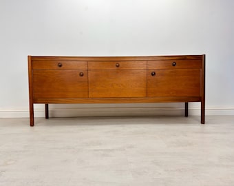 May include: A long, rectangular wooden sideboard featuring a warm brown finish. The piece includes three upper drawers and three lower cabinet doors, each equipped with small, round, dark brown wooden knobs. It stands on four straight legs.