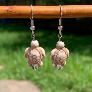 May include: A pair of silver dangle earrings featuring a white and brown turtle charm with a clear crystal bead at the bottom.