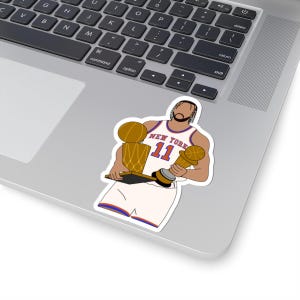 May include: A die-cut sticker featuring a stylized illustration of a basketball player wearing a white New York jersey with the number 11 in orange and blue. The player holds two gold-colored trophies, one in each hand, against a white background.