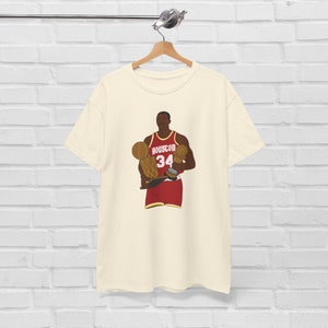 May include: A cream-colored t-shirt with a cartoon illustration of a basketball player holding three championship trophies. The player is wearing a red and white jersey with the number 34 and the word "HOUSTON" on the front.