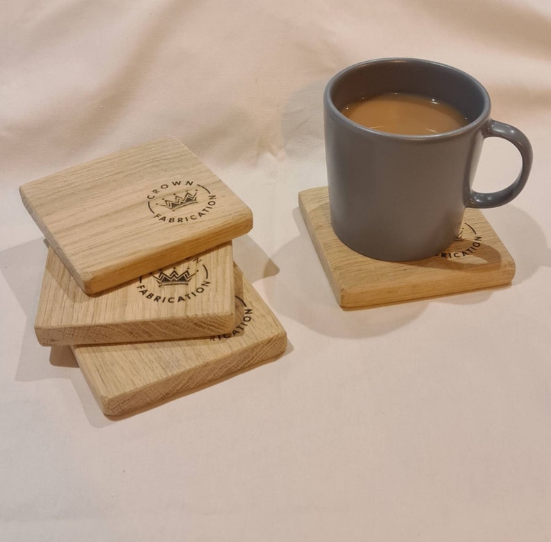 May include: A stack of four square wooden coasters with the text "CROWN FABRICATION" and a crown logo printed on them. A gray mug filled with a light brown liquid sits on a matching coaster. The coasters are made of light-colored wood.